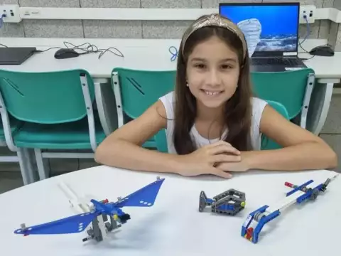 Menina sorridente com cabelos longos e faixa na cabeça sentada em uma sala de aula, interagindo com modelos de LEGO na mesa à sua frente.