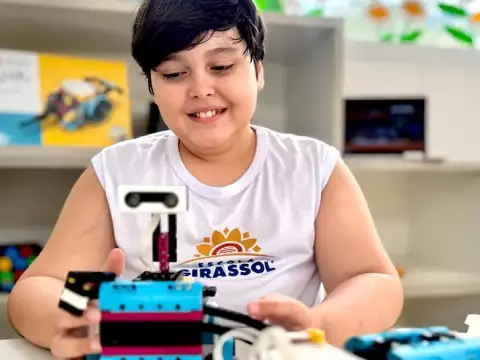 Uma criança de camisa branca sorri enquanto monta um robô com peças azuis e brancas sobre uma mesa em uma sala de aula.