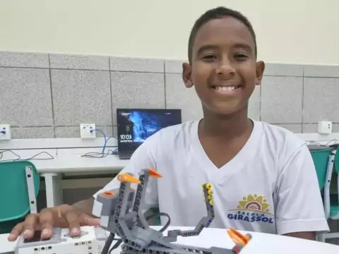 Uma criança de camisa branca sorri enquanto monta um robô com peças azuis e brancas sobre uma mesa em uma sala de aula.