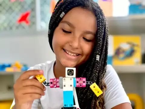Menina com tranças e pulseiras coloridas sorrindo enquanto ajusta um robô LEGO azul, branco e rosa com uma câmera na cabeça. O ambiente ao fundo contém prateleiras com materiais educativos e caixas de LEGO.